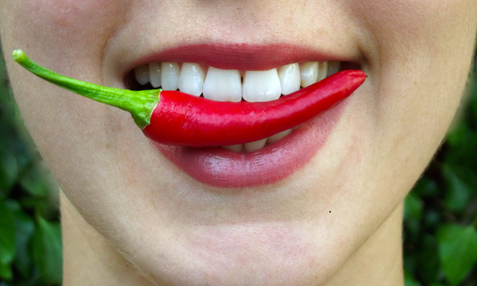 Close-up van een glimlachende mond met een vurige chilipeper tussen de tanden, die de pittigheid en sensatie van heet eten symboliseert.